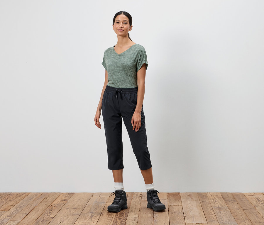 Frau steht vor einer weißen Wand und trägt ein grünes T-Shirt, schwarze Dreiviertelhosen und schwarze Wanderschuhe.