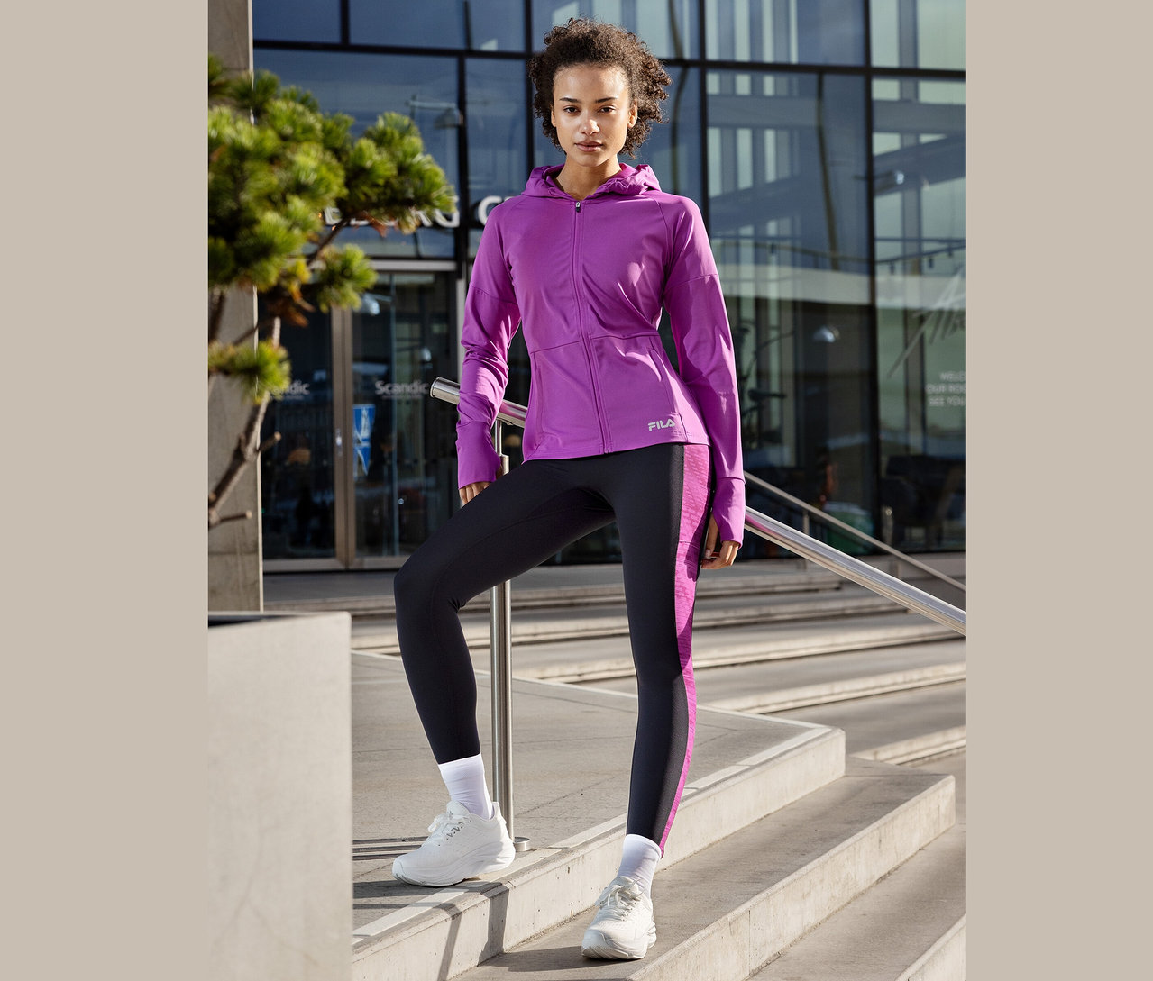 Frau posiert auf einer Treppe, trägt eine lila Fila-Jacke, Leggings mit lila Streifen und weiße Turnschuhe.