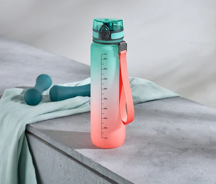 Eine Wasserflasche mit Farbverlauf steht neben zwei Hanteln auf einem Tuch.