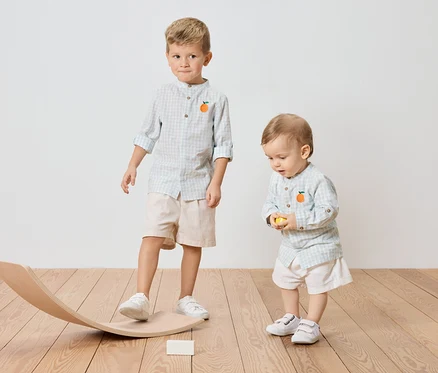 Zwei Kinder, ein älterer Junge, der auf einem Holzwippe steht, und ein kleiner Junge, der einen gelben Ball hält, beide tragen karierte Hemden mit Orangenapplikation.