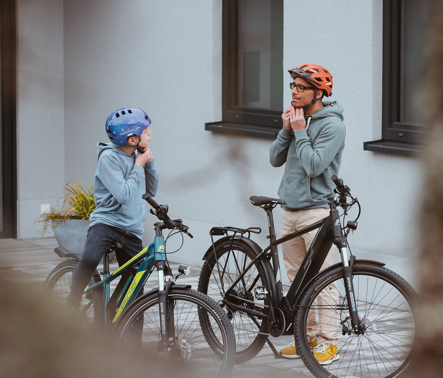 Ein Vater und ein Kind stehen vor ihren Fahrrädern und setzen ihre FISCHER Urban PLUS Miami Fahrradhelme auf.