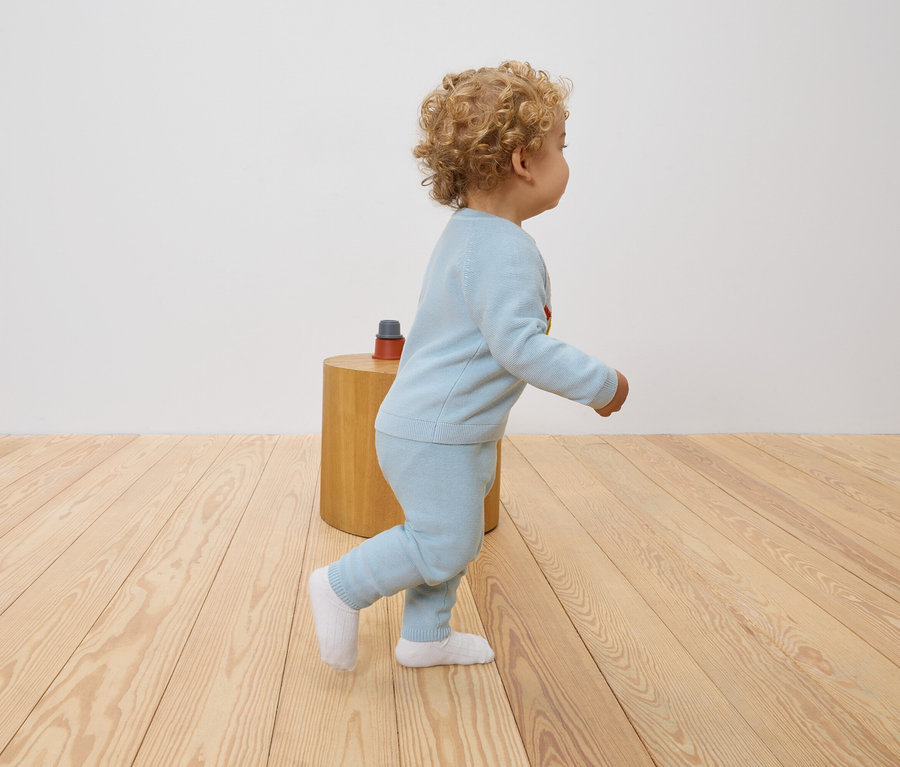 Ein Kleinkind mit lockigem Haar geht auf einem Holzboden. Es trägt einen hellblauen Pullover und passende Hosen.