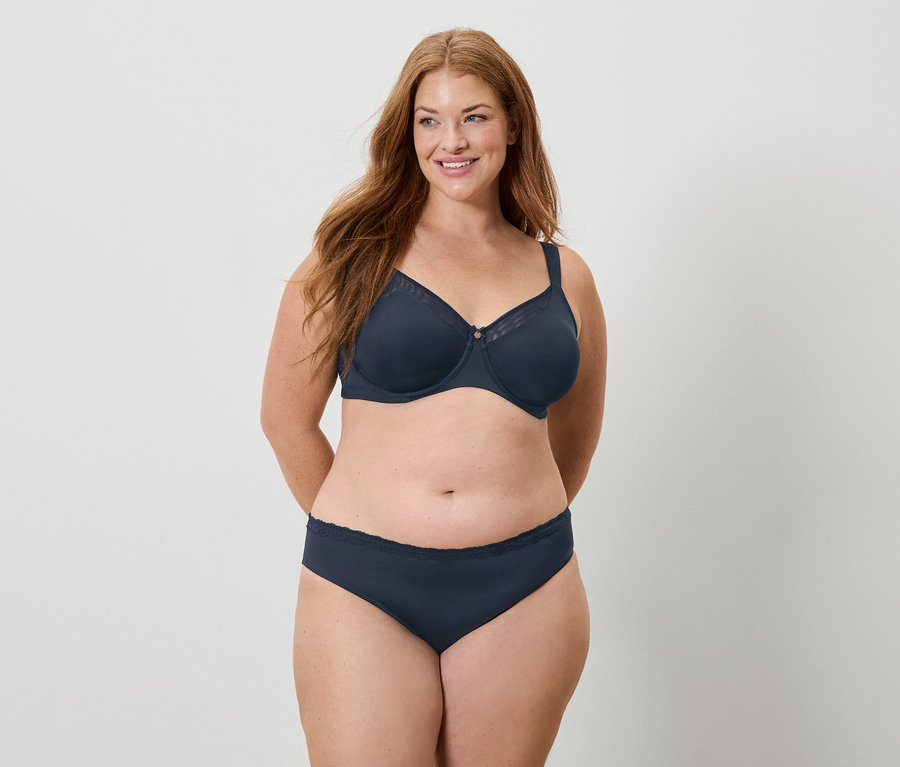 Frau mit langen roten Haaren trägt einen marineblauen BH und Slip und lächelt.