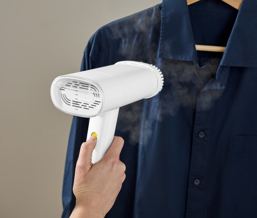 Eine Hand benutzt einen weißen Steamer, um ein blaues Hemd zu glätten, das an einem Holzbügel hängt.