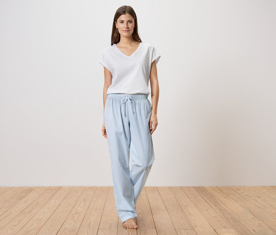 Eine Frau steht barfuß auf einem Holzboden und trägt ein weißes T-Shirt und eine hellblaue Pyjamahose.