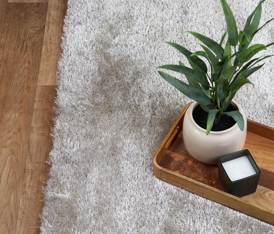 Nahaufnahme eines hellgrauen Teppichs »Silky« neben einem Holzboden. Auf dem Teppich steht ein Tablett mit einer Pflanze und einer Kerze.