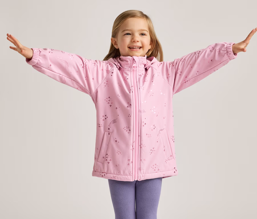 Ein lächelndes Mädchen mit blonden Haaren steht mit ausgestreckten Armen in einer rosa Jacke mit Sternen und lila Leggings vor einem hellen Hintergrund.