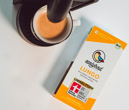 Eine Tasse Kaffee unter einer Kaffeemaschine und eine Packung Earlybird Lungo Kaffee sind auf einem Tisch zu sehen.