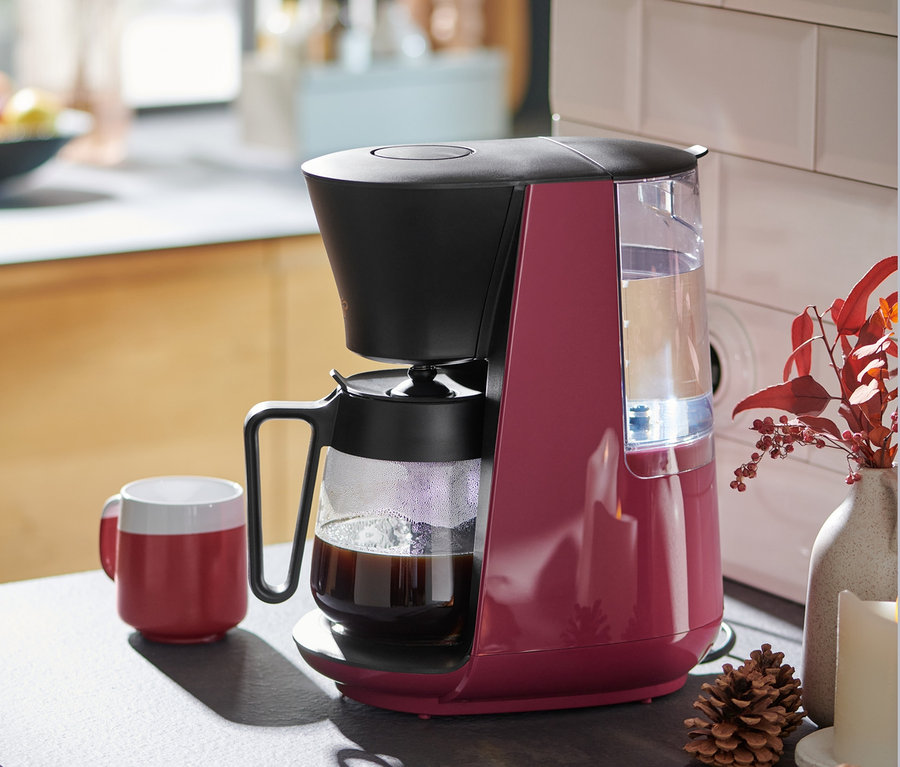 Beerenfarbene »Let's Brew« Filterkaffeemaschine steht neben einer roten Tasse Kaffee auf einer Küchentheke.