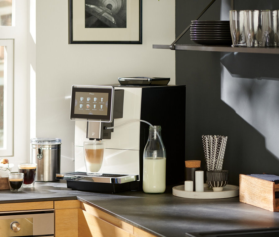 Auf einer Küchentheke steht ein weißer »Tchibo Office« Kaffeevollautomat, der Kaffee in ein Glas zubereitet. Daneben stehen eine Milchflasche, Croissants, Kaffee und ein Notizbuch.