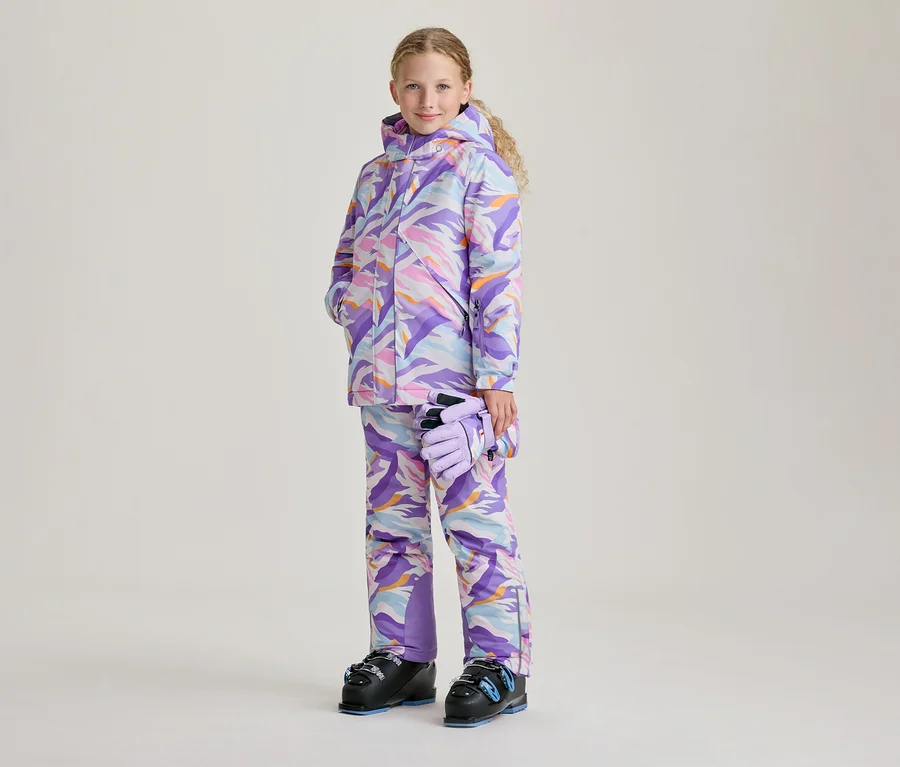 Ein junges Mädchen mit blonden Locken steht lächelnd in einem bunten Skianzug mit abstraktem Muster in Lila, Pink, Blau und Orange. Sie hält hellviolette Handschuhe in der linken Hand und trägt schwarze Skischuhe mit blauen Akzenten.