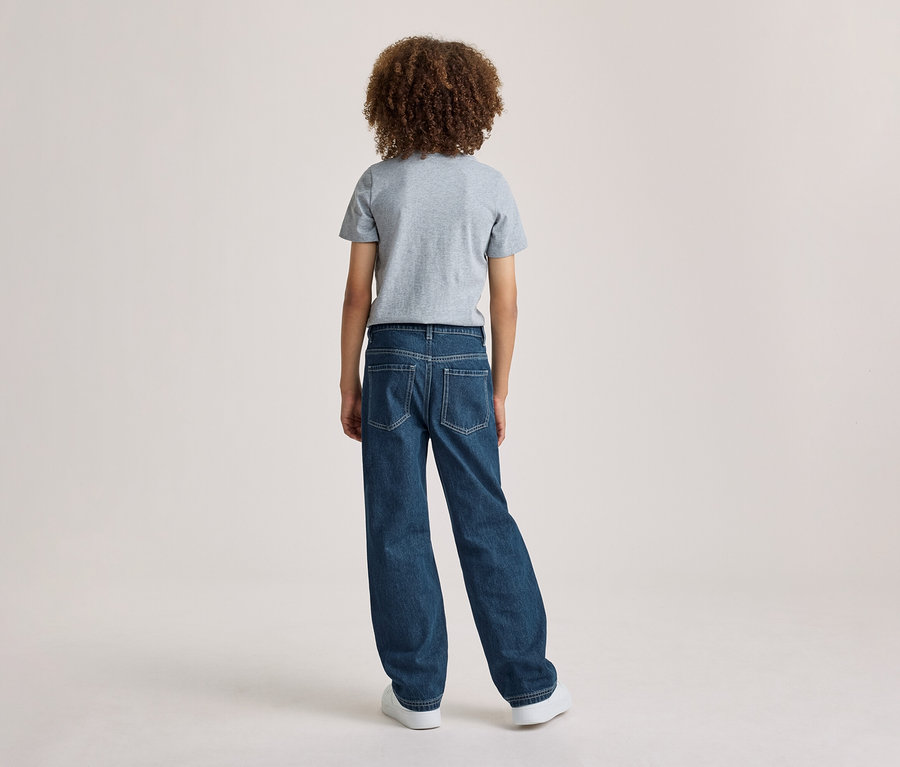 Eine Person mit lockigem Haar, von hinten gesehen, trägt ein graues T-Shirt und blaue Jeans.
