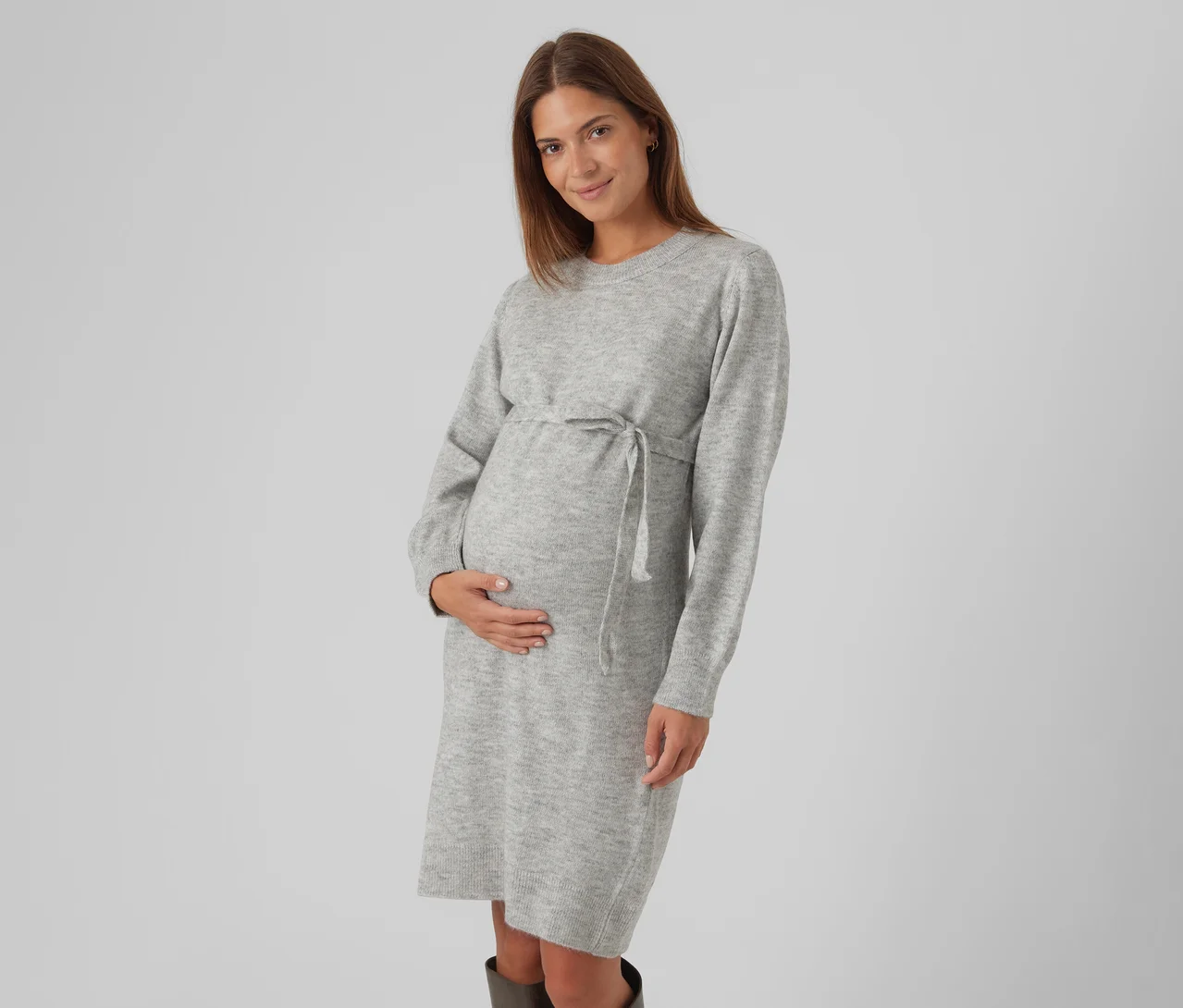 Schwangere Frau mit langen braunen Haaren, die ein hellgraues Strickkleid mit Kordelzug an der Taille trägt, ihren Babybauch sanft hält und lächelt.