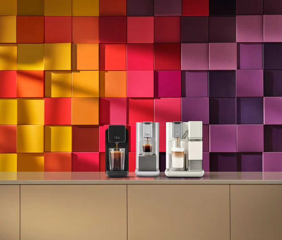 Drei Qbo Kaffeemaschinen auf einer beigen Arbeitsplatte. Eine schwarze Maschine gießt Espresso in ein Glas, eine silberne Maschine gießt Espresso in ein weiteres Glas und eine weiße Maschine mit Milchaufschäumer bereitet einen Latte zu. Dahinter befindet sich eine Wand mit 3D-Quadratfliesen in einem Farbverlauf von Gelb zu Violett.