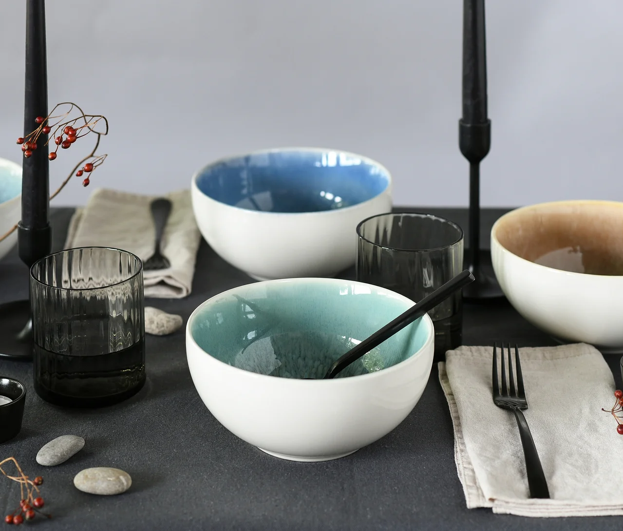 Tisch gedeckt mit drei weißen creaTable Caldera Schalen mit blauer, türkiser und hellbrauner Innenseite, dunkelgrauen Gläsern, schwarzem Besteck und schwarzen Kerzen auf einer dunkelgrauen Tischdecke.
