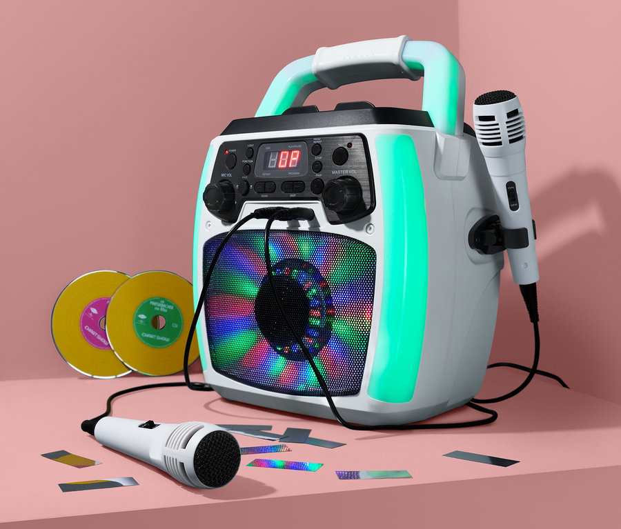 Ein weißes Karaoke-System mit zwei Mikrofonen steht auf einer pinkfarbenen Oberfläche neben zwei CDs und Konfetti.