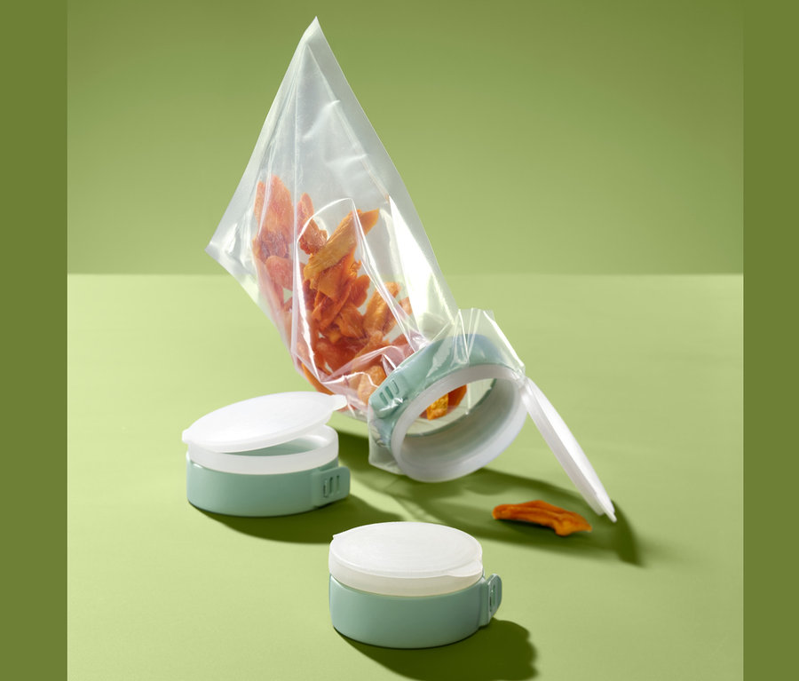 Eine durchsichtige Tüte mit orangefarbenen Essensstücken wird mit 3 Aroma-Tütenverschlüssen in Grün verschlossen. Zwei weitere Verschlüsse liegen daneben.