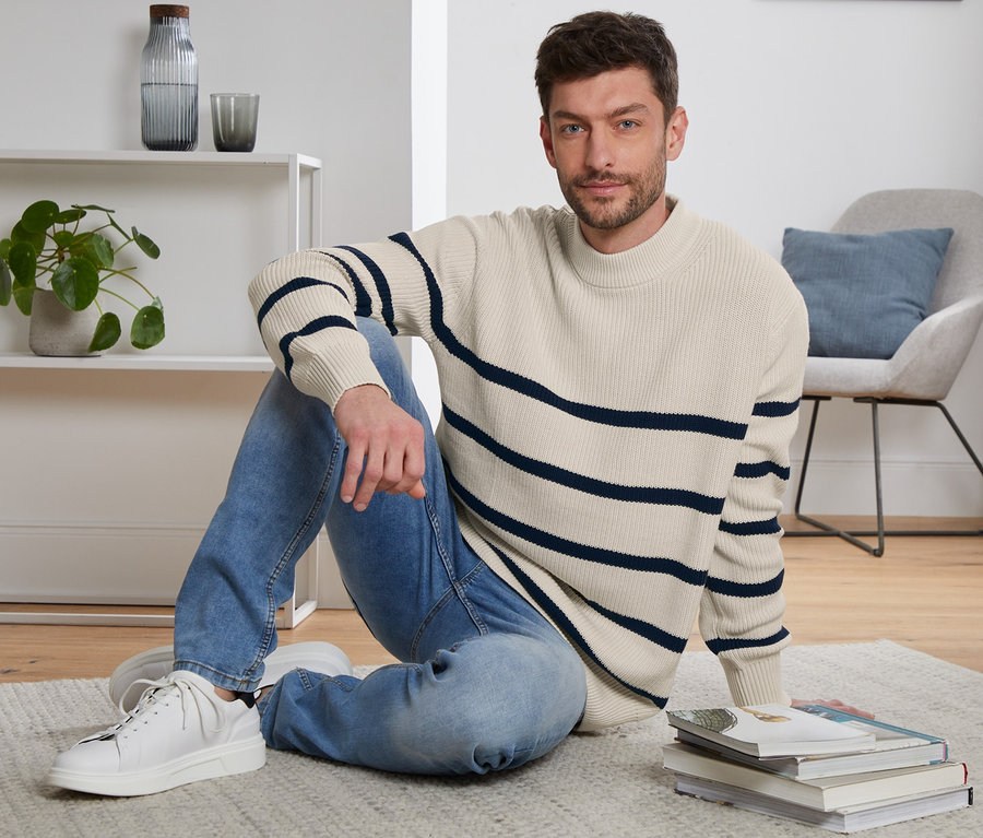 Mann sitzt auf dem Teppichboden und trägt einen Strickpullover.