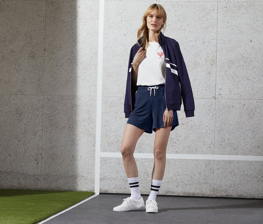 Eine Frau steht und trägt dunkelblaue Jerseyshorts, ein weißes T-Shirt und eine blaue Jacke.