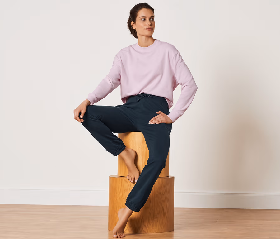 Frau sitzt auf Holzblöcken und trägt eine dunkelblaue Sweathose und ein rosa Sweatshirt.