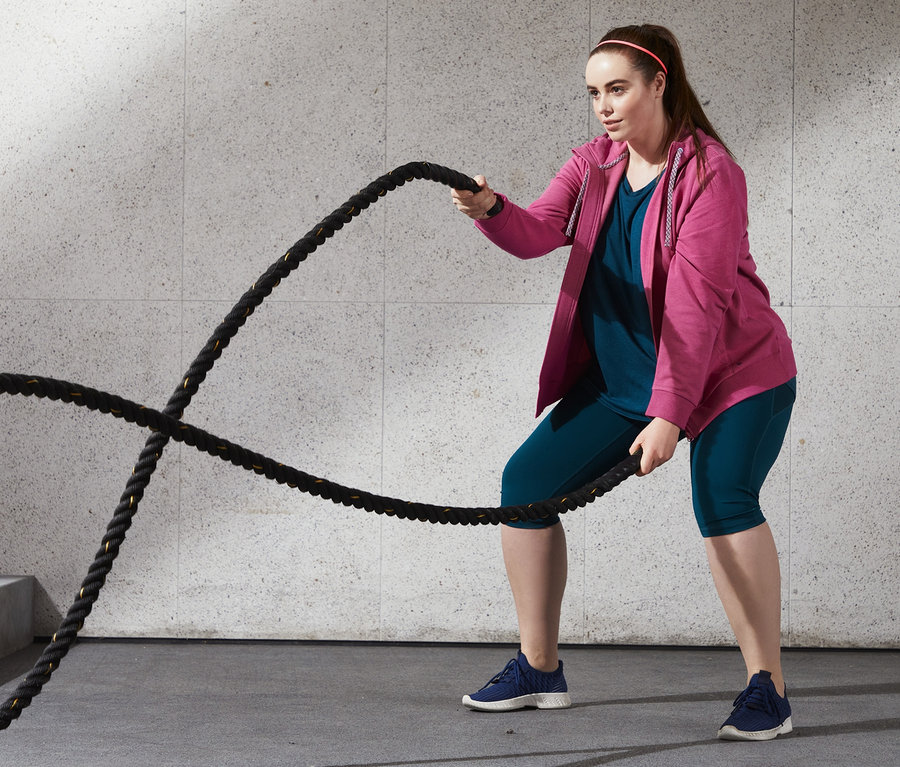 Frau trainiert mit Seilen in roséfarbener melierter Sweatjacke mit Kapuze, 3/4-Sport-Tight und blauem Sportshirt.