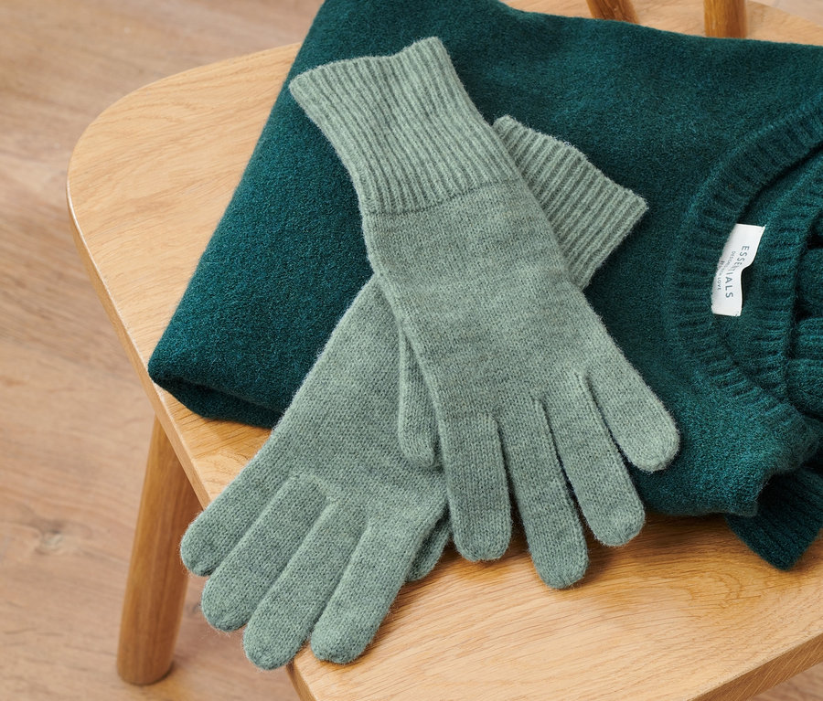 Auf einem Holzstuhl liegen ein grüner Pullover und Strickhandschuhe mit Wolle in Salbeigrün.
