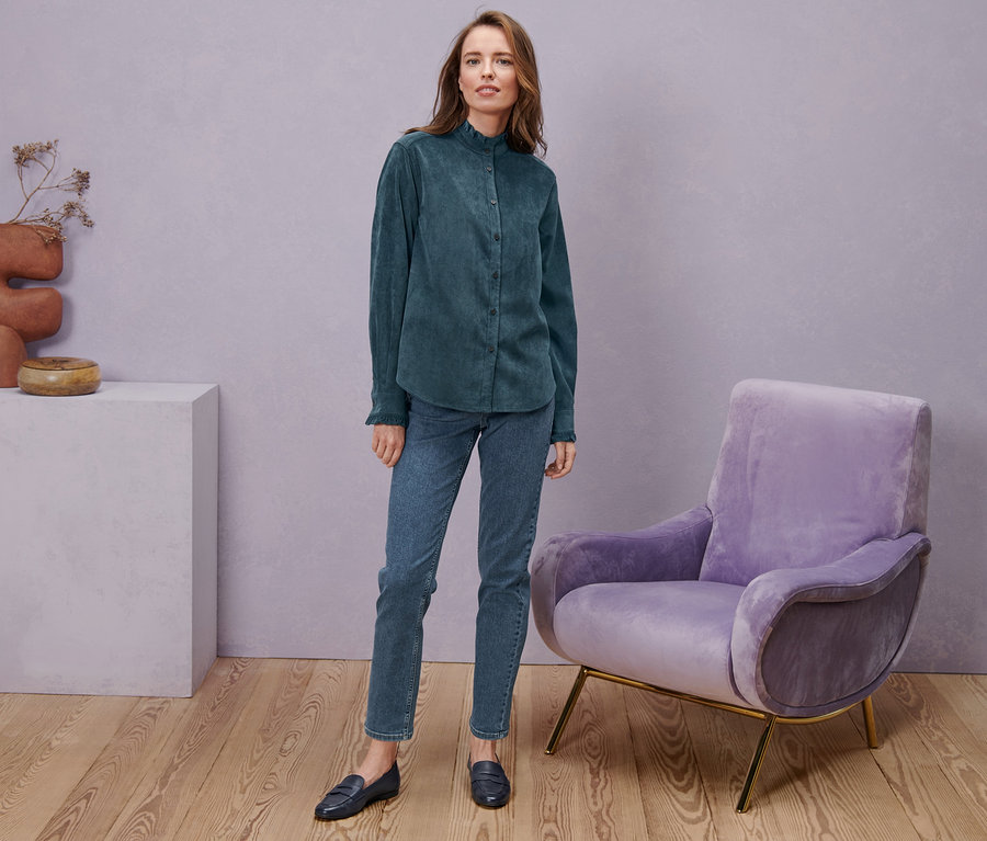 Eine Frau steht und trägt Straight-Denim »Fit Juna«, midblue, eine Cordbluse mit Rüschendetail und Leder-Loafer.