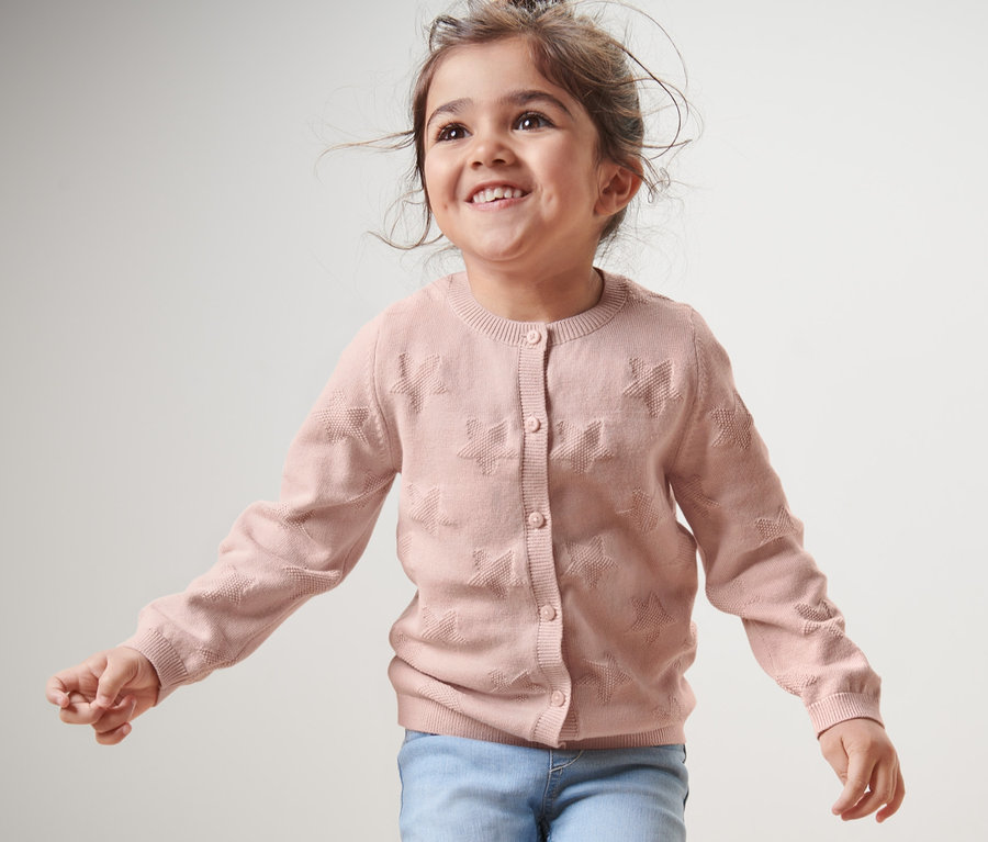 Lächelndes Mädchen trägt eine Kinder-Strickjacke und Jeans.