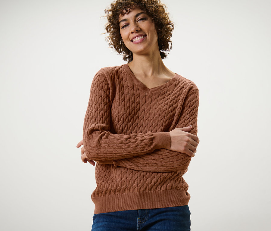 Lächelnde Frau posiert mit verschränkten Armen in einem cognacfarbenen Zopfstrickpullover und Jeans.