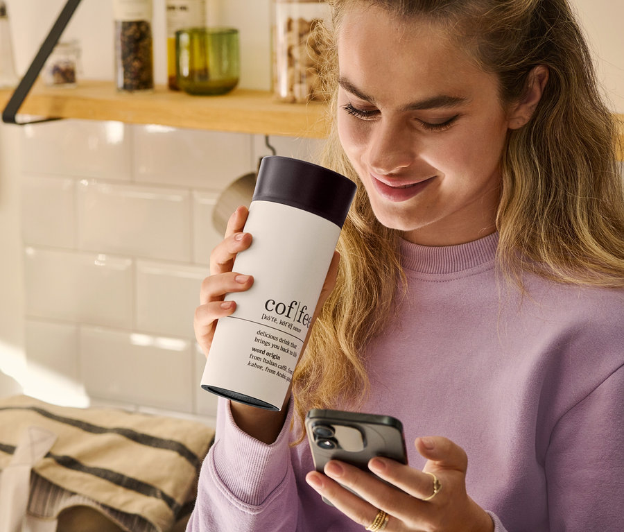 Frau hält einen Coffee-to-go-Becher, Coffee Dictionary, in der Hand und schaut auf ihr Mobiltelefon.