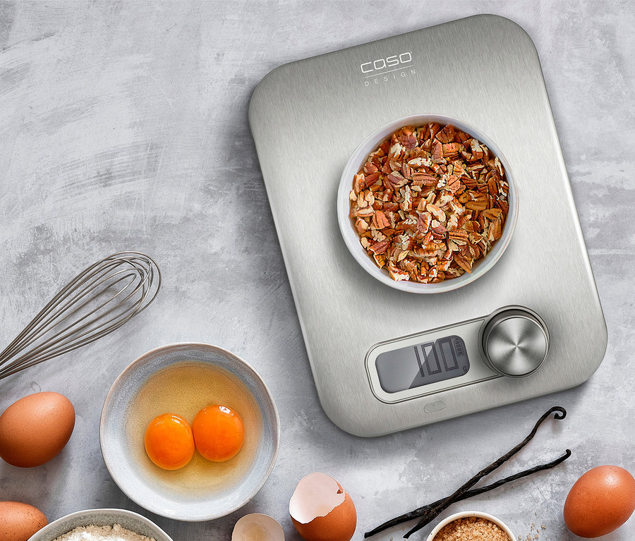 CASO Küchenwaage »Kitchen Energy« mit Nüssen in einer Schüssel, Eiern, Mehl, Zucker und Honig auf grauem Hintergrund.