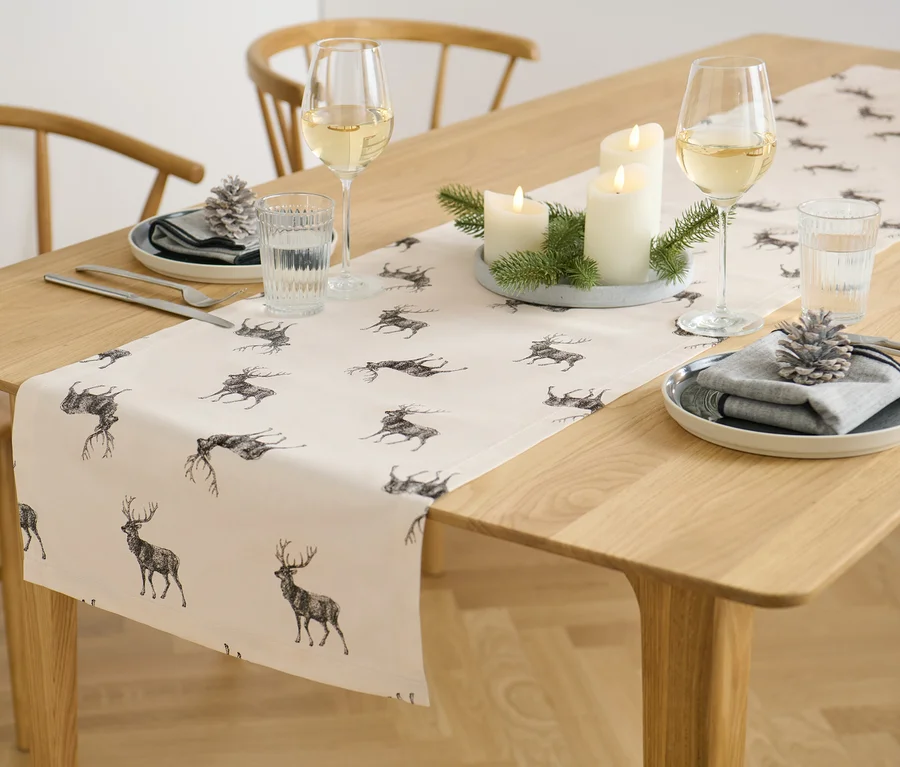 Auf einem Holztisch liegen ein Tischläufer mit Hirsch-Alloverprint, daneben Kerzen, Weingläser und Teller.