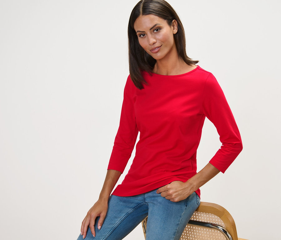 Frau sitzt auf einem Stuhl und trägt ein rotes Shirt mit 3/4-Arm.