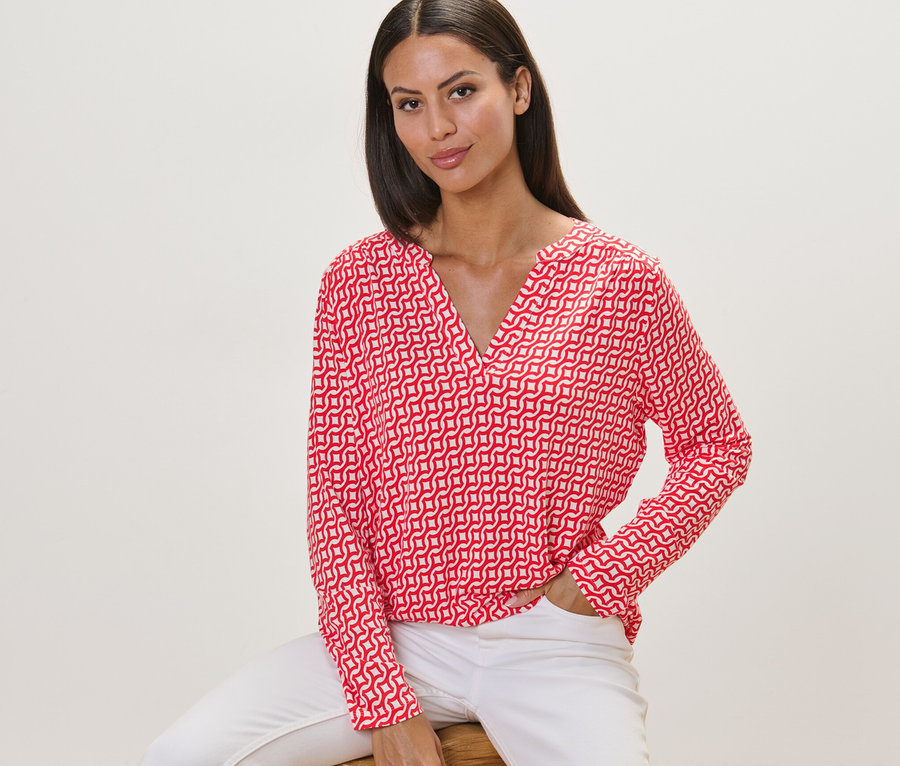 Frau sitzt auf einem Hocker und trägt ein bedrucktes Blusenshirt in Rot.