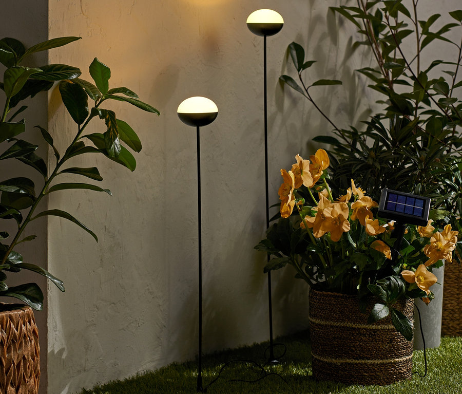 Zwei Solar-Leuchtstäbe stehen auf Rasen zwischen Blumen in Töpfen.