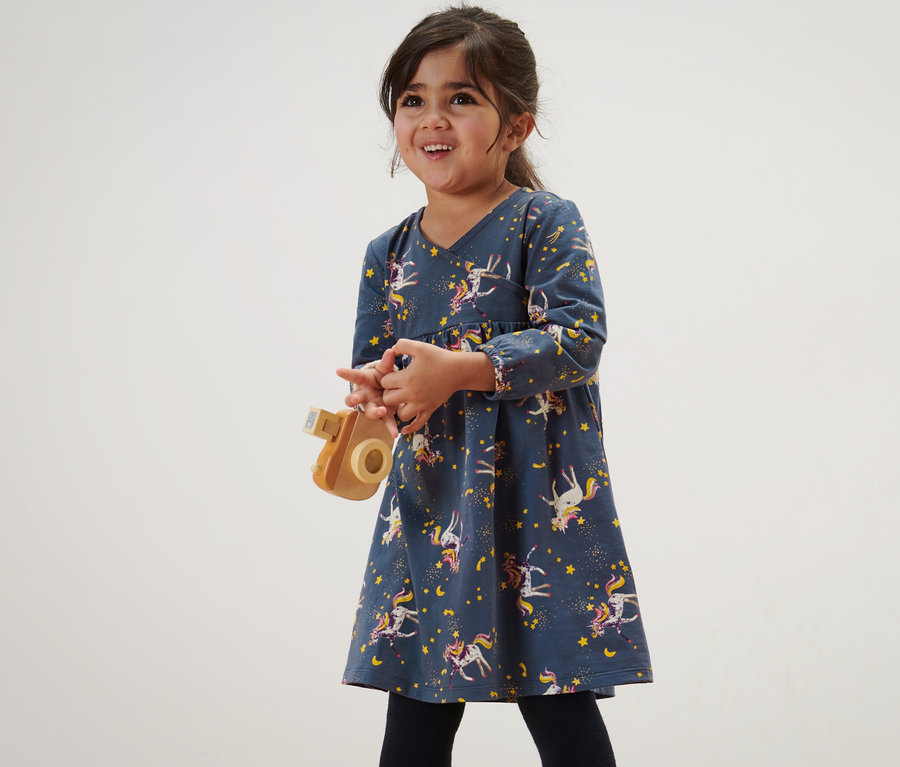 Lächelndes Mädchen hält eine Holzkamera und trägt ein Kinder-Jerseykleid mit Einhornmuster.