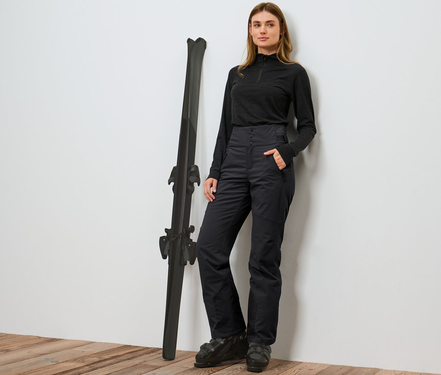 Frau posiert neben Skiern in einer schwarzen High-Waist-Skihose.