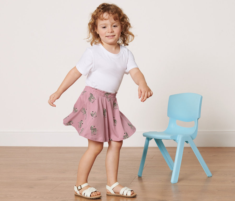 Mädchen in weißem T-Shirt und Kinder-Jerseyrock mit Hose steht neben blauem Stuhl.