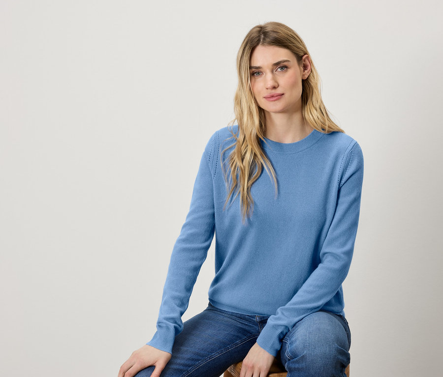 Frau sitzt auf einem Hocker und trägt einen blauen Pullover und Jeans.