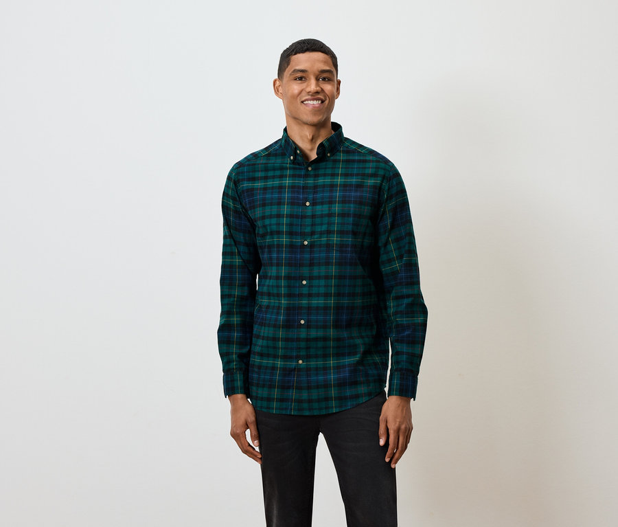 Ein kurzhaariger Mann posiert vor einem weißen Hintergrund und trägt ein grün-blau kariertes Langarmhemd und schwarze Jeans.