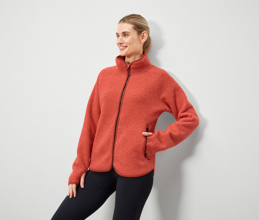 Frau posiert in orangefarbener Fleecejacke mit Reißverschluss und schwarzen Leggings.