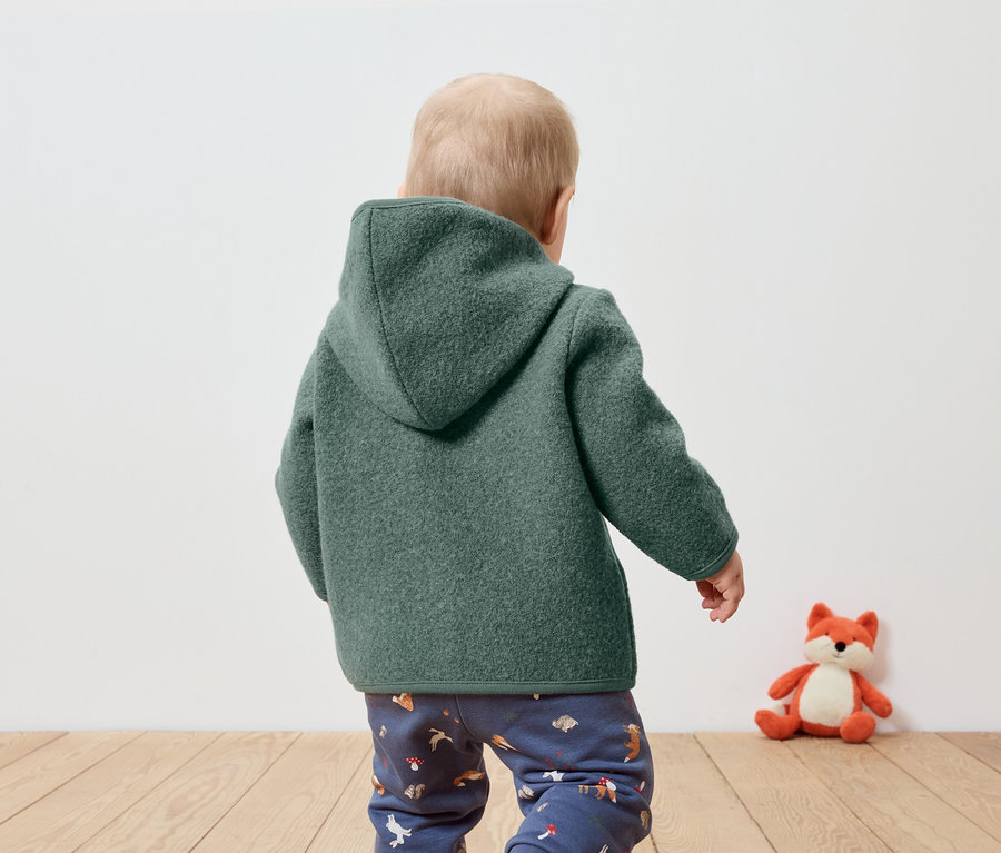 Kleinkind mit dem Rücken zur Kamera trägt einen grünen Kapuzenmantel und gemusterte Hosen, daneben sitzt ein Plüschfuchs.