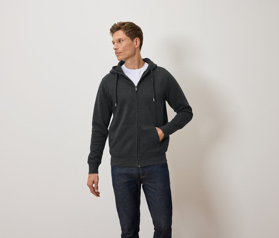 Mann trägt einen dunkelgrauen Kapuzenpullover mit Reißverschluss, ein weißes T-Shirt und blaue Jeans.