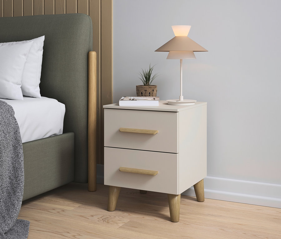 Ein Nachttisch mit zwei Schubladen steht neben einem Bett, auf dem Tisch stehen eine Lampe, ein Buch und eine kleine Pflanze.