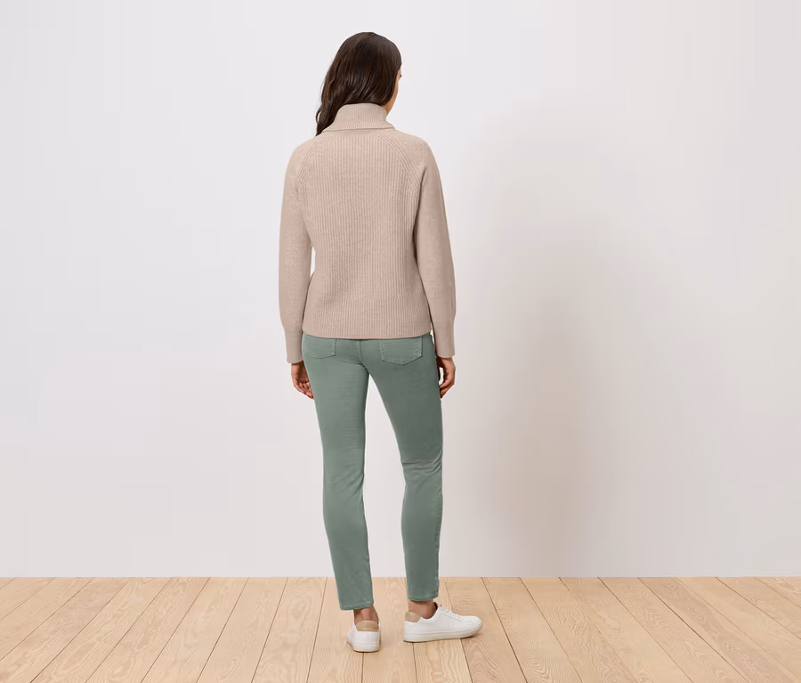 Eine Frau steht mit dem Rücken zur Kamera und trägt einen beigen Rollkragenpullover, grüne Jeans und weiße Turnschuhe.