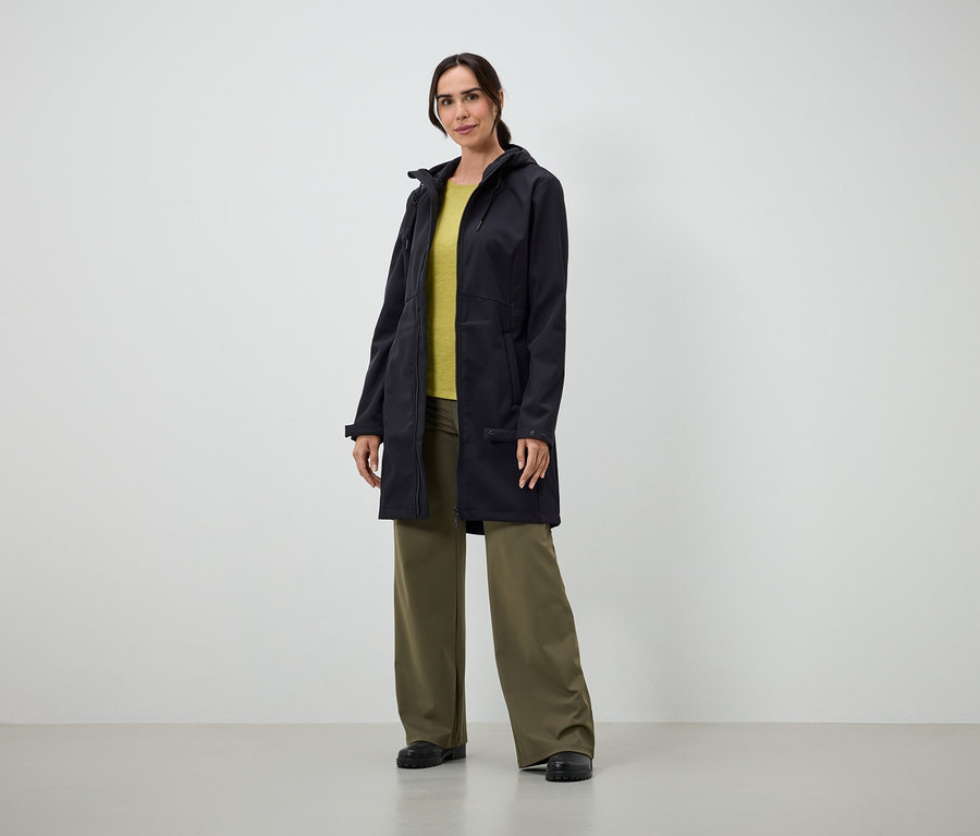 Frau in langem schwarzen Mantel, gelbem T-Shirt und weiter grüner Hose steht drinnen.