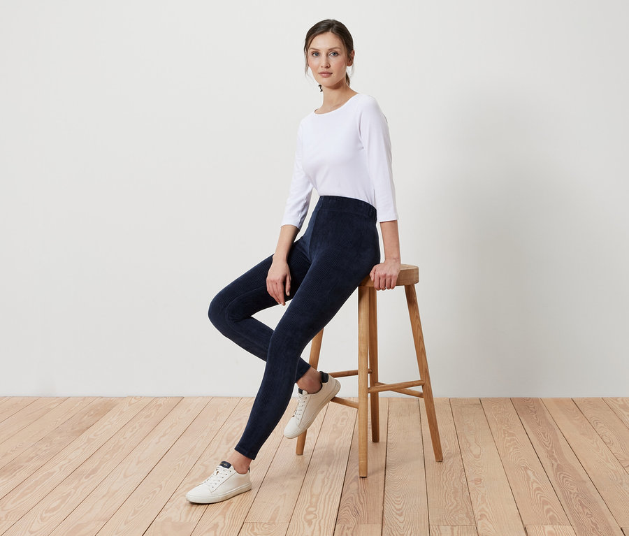 Eine Frau sitzt auf einem Holzhocker und trägt marineblaue Leggings und ein weißes Shirt.