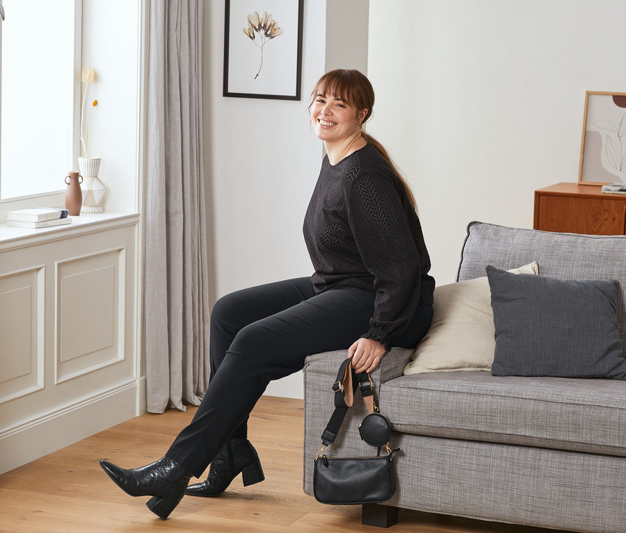 Frau sitzt auf Sofa. Sie trägt Leder-Boots, Stretchhose, dunkelrot und hält Mini-Tasche, rot.