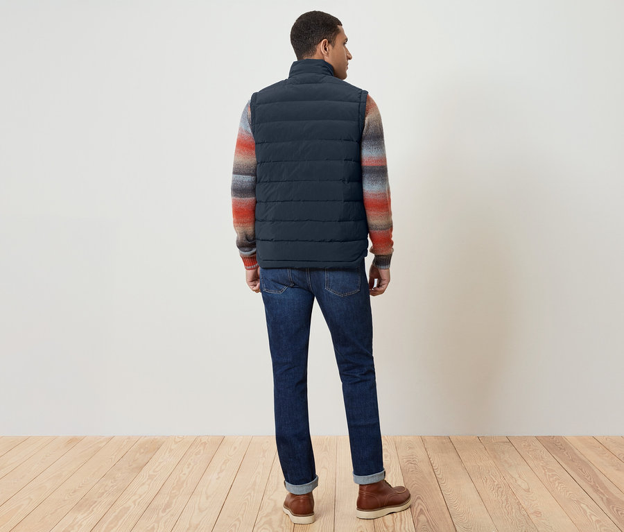 Ein Mann mit dem Rücken zur Kamera trägt eine marineblaue Weste, einen bunten Pullover, Jeans und braune Schuhe.