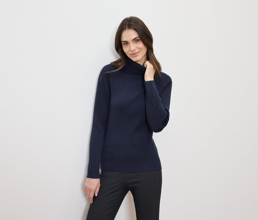 Frau mit braunen Haaren posiert in einem dunkelblauen Rollkragenpullover und einer schwarzen Hose.